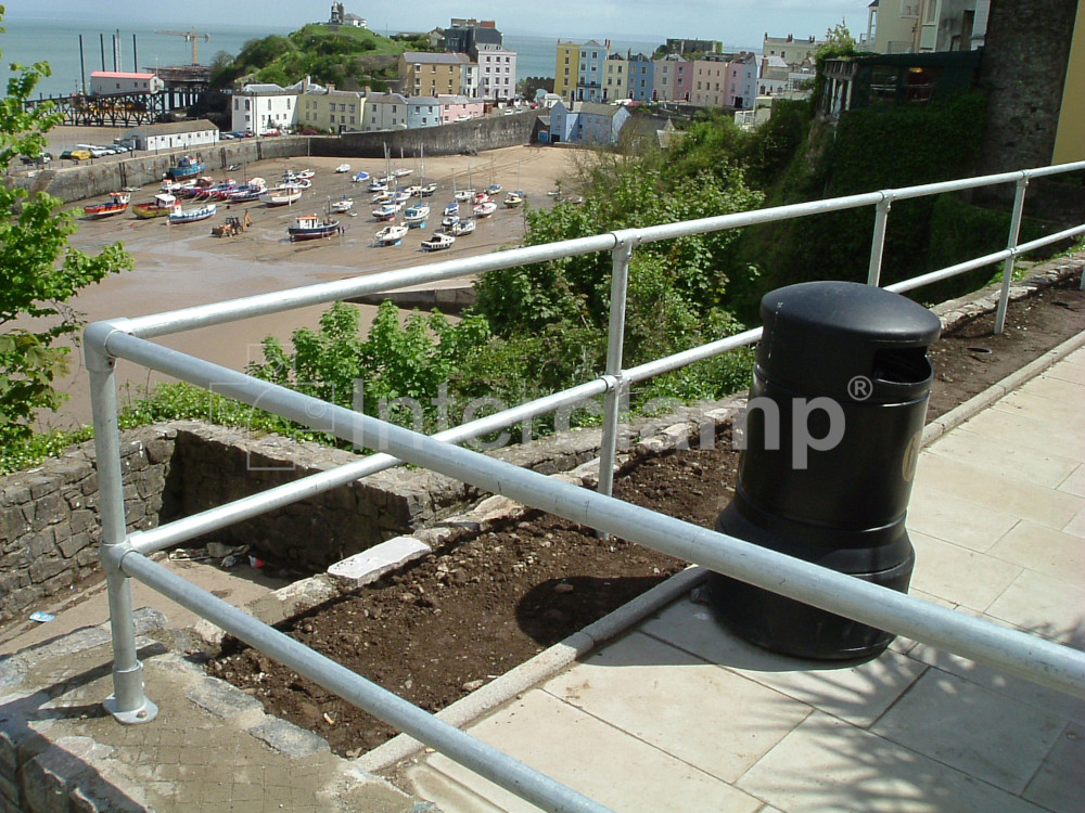 A seaside town promenade featuring a robust Interclamp tube clamp handrail, with people enjoying a stroll and the beach visible in the distance.